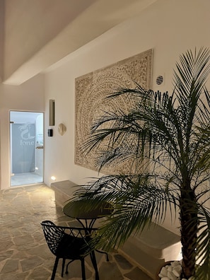 A modern interior with a stone floor, a black chair, a large wall art piece, a potted plant, and a door leading to a bathroom. - Ione Inn (Chora)
