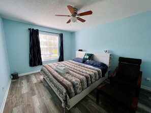 Ein Schlafzimmer mit Bett, Deckenventilator, Fenster mit Vorhängen und einem Sessel.