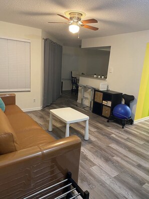 Ein Wohnzimmer mit braunem Ledersofa, einem weißen Couchtisch, Deckenventilator und einer Küchenzeile mit einer Theke und einem Fitball.