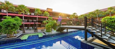 Une piscine dotée d’une terrasse carrelée, entourée de palmiers et bordée d’un bâtiment au toit rouge.