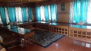 A room with a wooden bench, a glass-top table, patterned carpet, and blue curtains. - Hotel Chubi Leh (Leh)