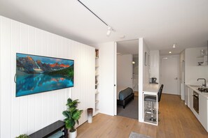 Ein modernes Interieur mit einem an einer weißen Wand montierten Flachbildfernseher, einer Essecke mit einer Bank und Stühlen sowie einer Küche mit weißen Schränken und einer Arbeitsplatte.
