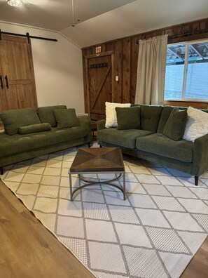Una habitación con dos sofás verdes, una mesa de centro de madera, una alfombra estampada, una puerta de madera con el rótulo 'EXIT' y una ventana con cortinas.