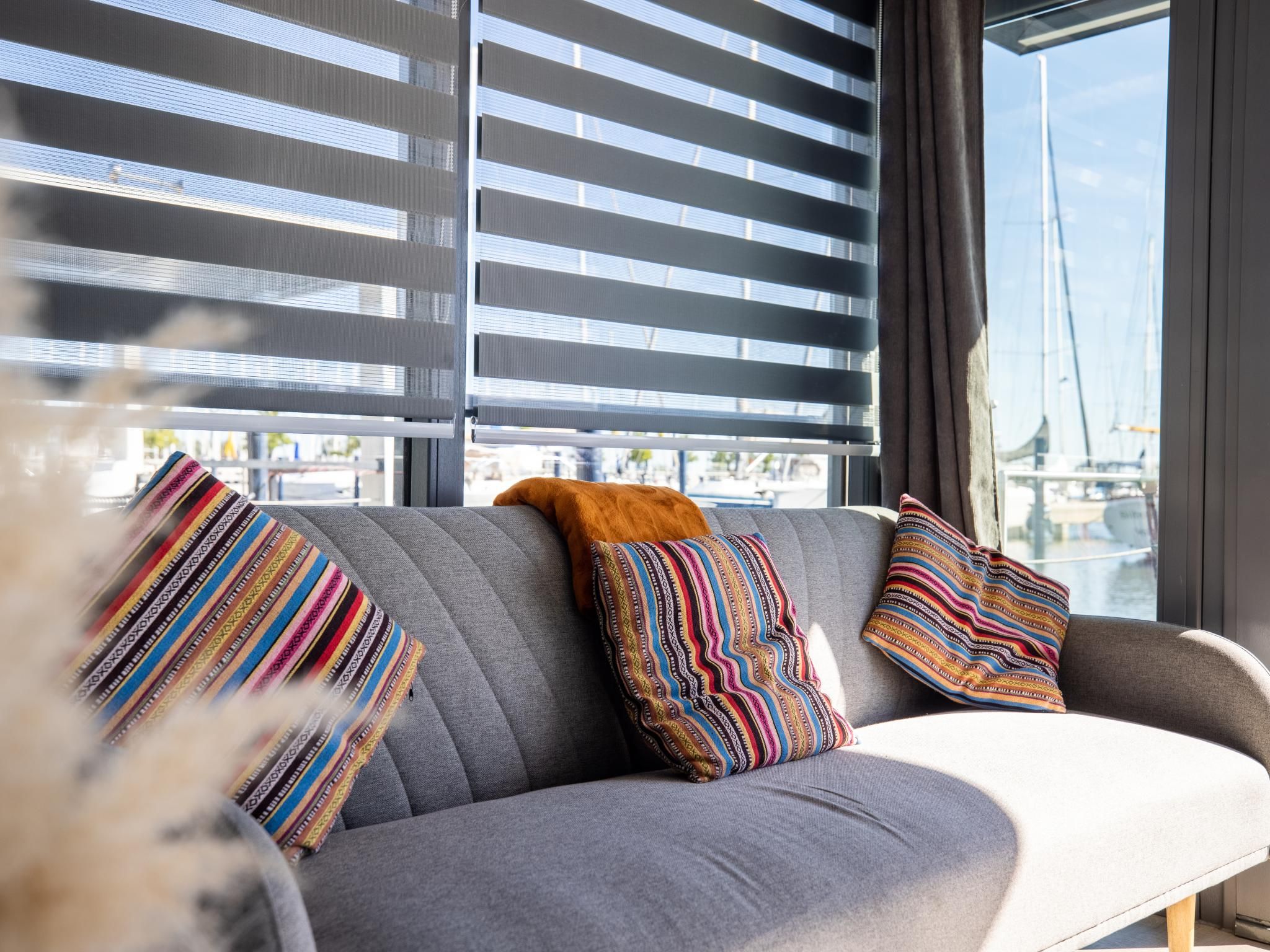 A modern living room with a grey sofa, striped pillows, and a view of a cityscape through large windows with blinds.