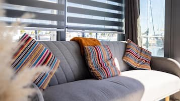 A modern living room with a grey sofa, striped pillows, and a view of a cityscape through large windows with blinds.