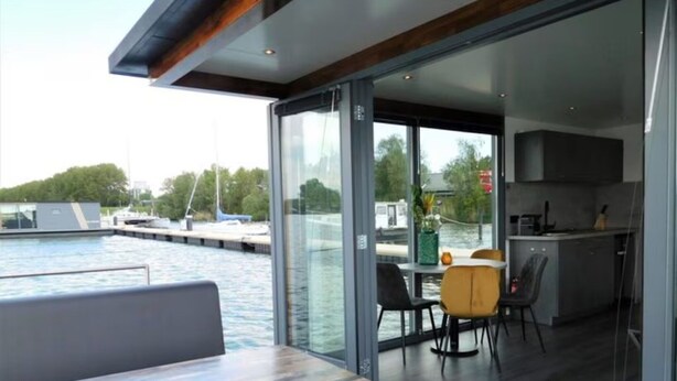 Un interior de barco moderno con comedor, cocina y grandes ventanales con vistas al agua y al muelle.