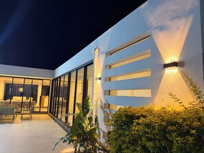 Fachada moderna de edifício com grandes portas de vidro, área de estar com cadeiras e um painel de parede bem iluminado.