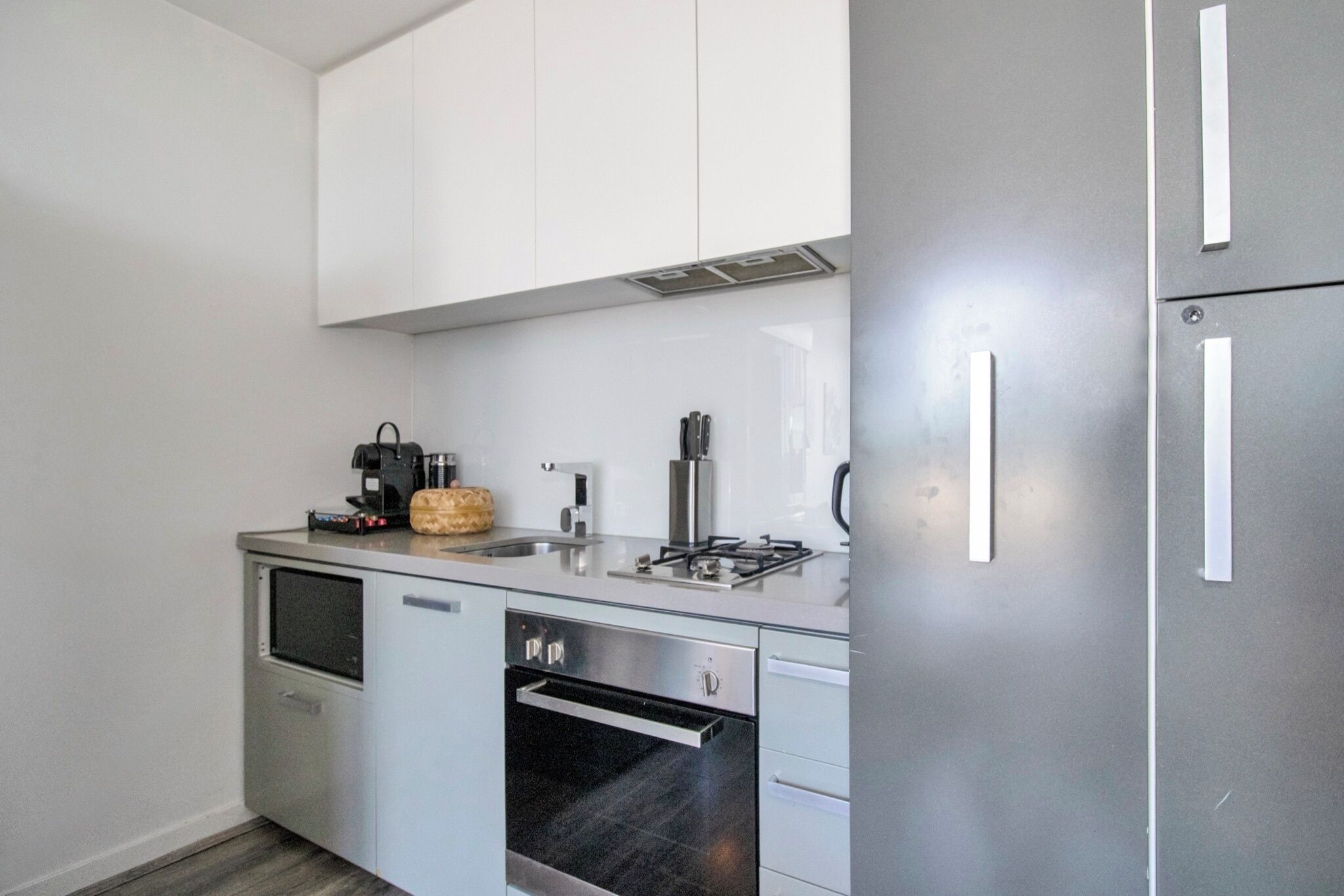 A compact kitchen with white cabinets, a stainless steel refrigerator, a built-in oven, and a coffee maker.