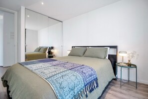 A modern bedroom with a large bed, a bedside table with a lamp, and a mirror on the wall. - Top-Floor Apartment with Balcony - High Street (Prahran)