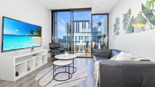 A modern living room with a flat-screen TV, a sofa, a coffee table, and a balcony with a city view. - Top-Floor Apartment with Balcony - High Street (Prahran)