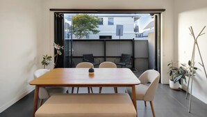 Een moderne eetruimte met een houten tafel en stoelen, een bank, en uitzicht door een schuifdeur op een buitenruimte met een boom en gebouwen.