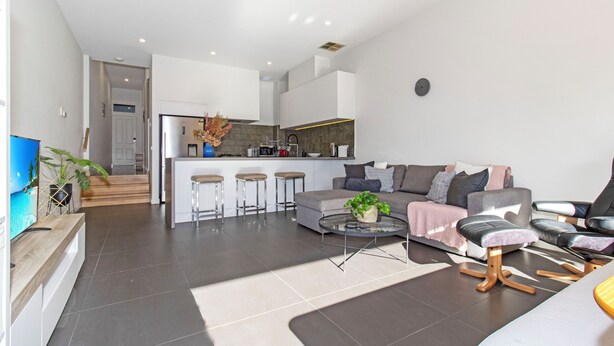 Een moderne woonkamer met een bank, salontafel en barstoelen. Achtergrond toont een keuken.