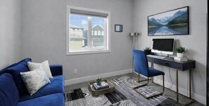 Ein Wohnzimmer mit einem blauen Sofa, einem Schreibtisch mit Computer, einem Fenster mit Blick auf Häuser und einem gerahmten Bild an der Wand.