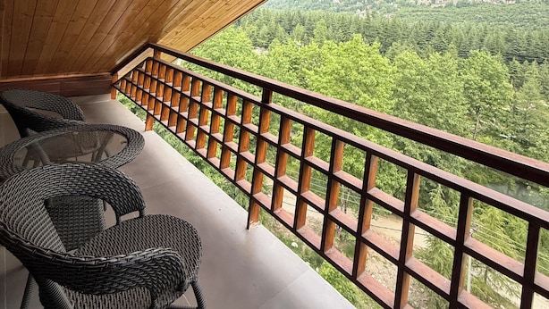 A wooden balcony with wicker chairs, a railing, and a view of a forested valley. - Old Manali Retreat (Samudrapur)