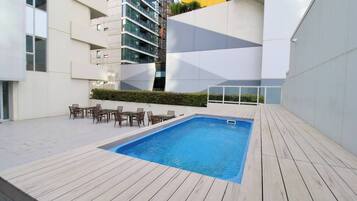 A rooftop pool with wooden decking, surrounded by modern buildings.