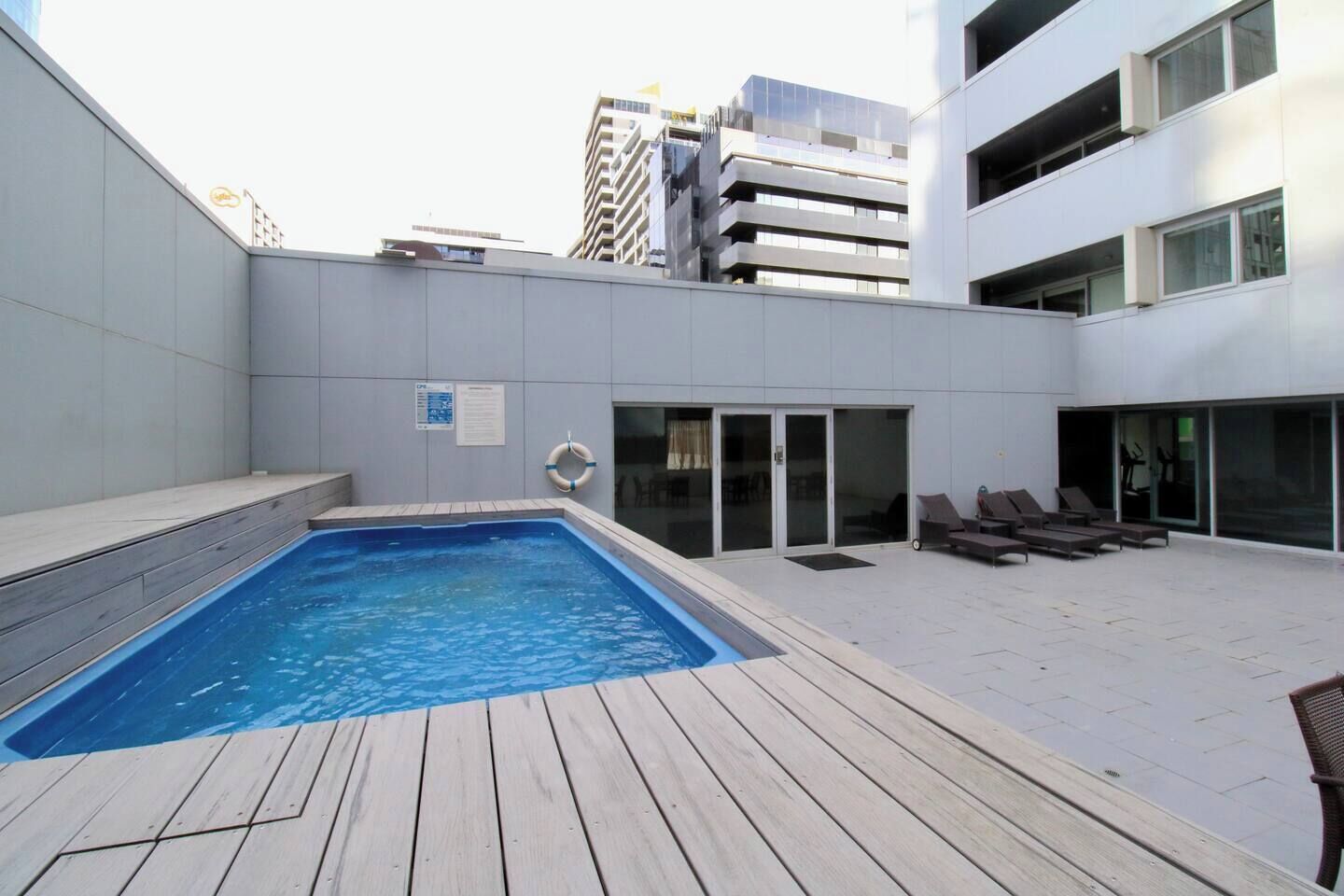 Uma área de piscina no terraço com deck de madeira, cadeiras de descanso e entrada por porta de vidro.