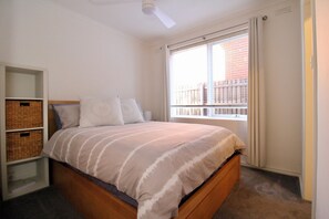 A bedroom with a bed, a window with a view of buildings, a ceiling fan, and built-in storage. - Park Street Apartment - St Kilda West (Saint Kilda West)