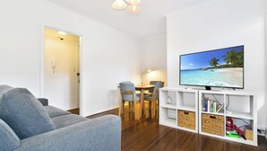 Ein Wohnzimmer mit grauem Sofa, einem Esstisch mit Stühlen, einem Fernseher auf einem weißen Schrank und einem Bücherregal.