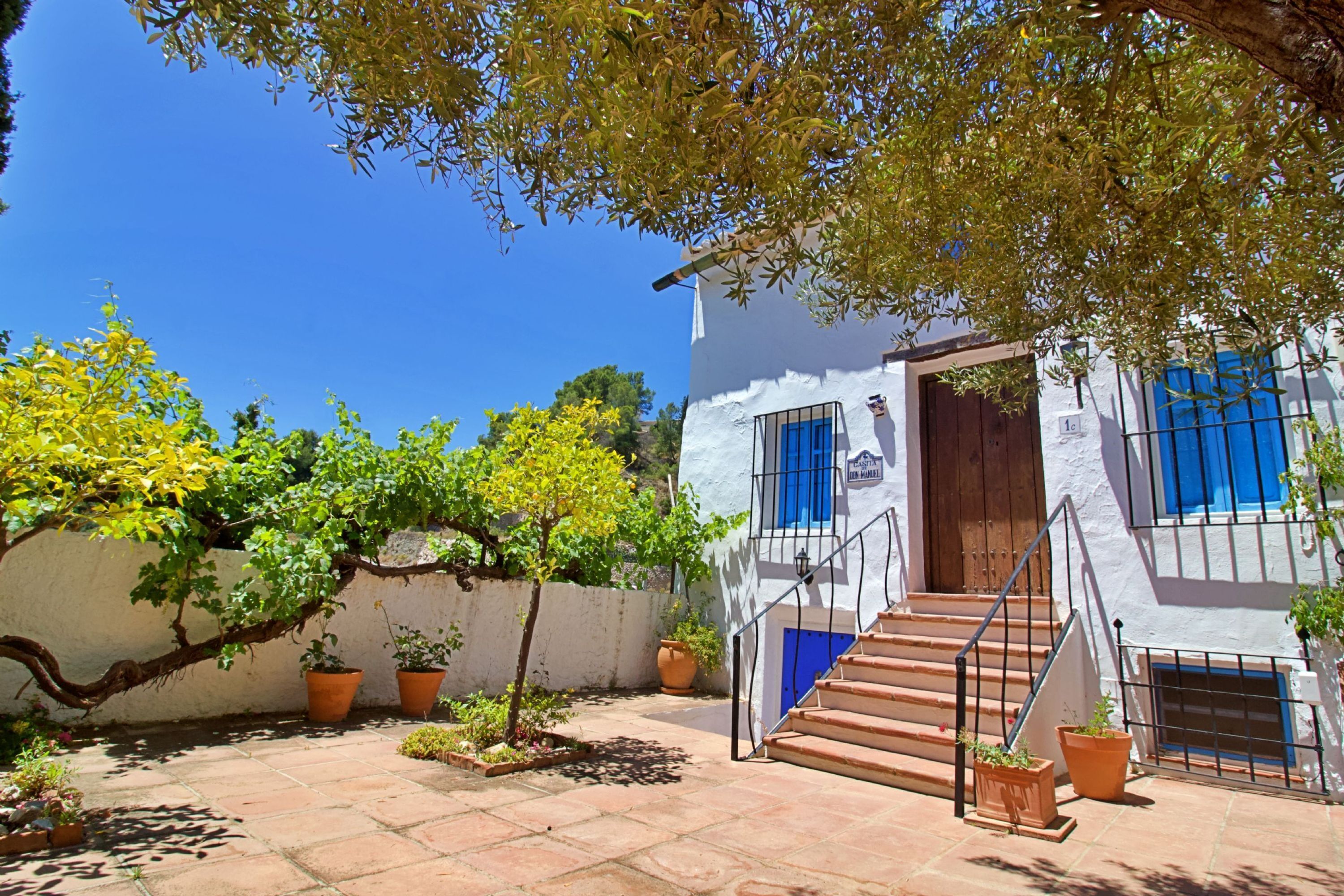 A white building with blue shutters, a wooden door, and a staircase leading to the entrance. There are potted plants and a tree in the courtyard.