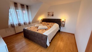 Quarto com cama, cortinas, cabeceira, luminárias fixadas na parede, radiador e um quadro.