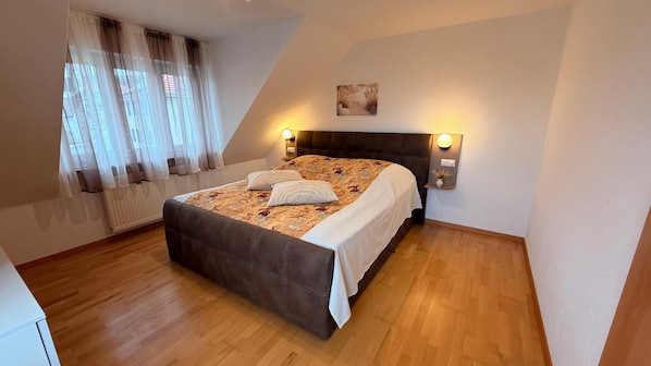 Un dormitorio con cama, cortinas, cabecera, luces fijadas en la pared, radiador y un cuadro.
