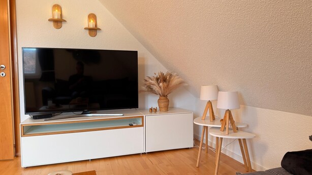 Uma sala de estar moderna com televisão de tela plana, armário de madeira, luminárias decorativas e um vaso com plumas de capim-do-campo.