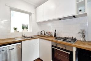 A modern kitchen with white cabinets, a dishwasher, a stove, and a window with plants. - Beachfront Marine Parade - Rooftop BBQ - Luna Park (Saint Kilda, Victoria, AU)