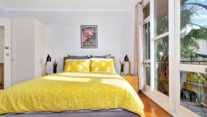 A bedroom with a large bed, a poster on the wall, and a view of palm trees through the window. - Beachfront Marine Parade - Rooftop BBQ - Luna Park (Saint Kilda, Victoria, AU)