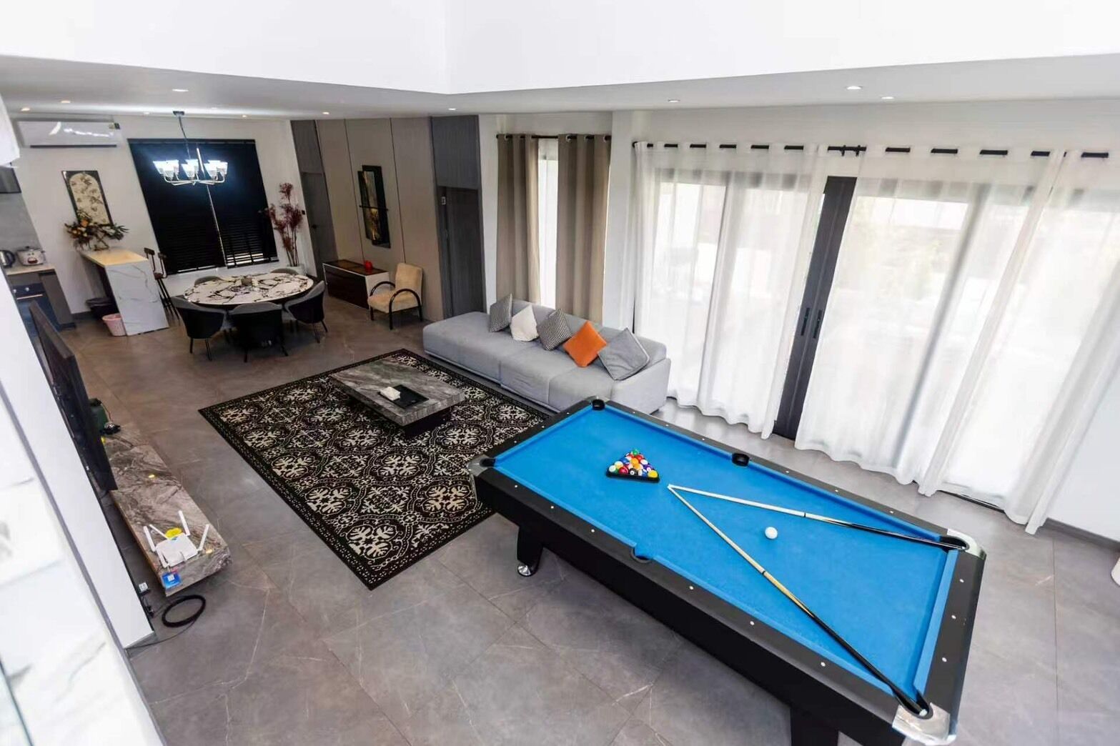 Een moderne woonkamer met biljart, een bank, een eethoek en een groot raam met gordijnen.