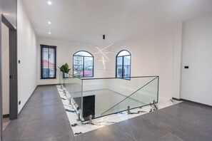 A modern interior with a glass railing, a geometric light fixture, and a view through large windows. - Acadia 5BR (Banglamung)
