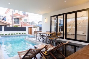 A modern outdoor pool area with a dining table and chairs, a lounge chair, and a view of the surrounding buildings. - Acadia 5BR (Banglamung)