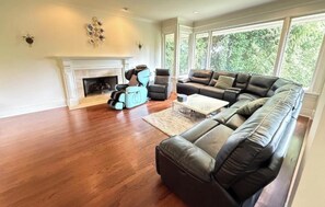 A spacious living room with a fireplace, leather sofas, and a large window offering a view of greenery. - Luxury Accommodation in West Van (West Vancouver)