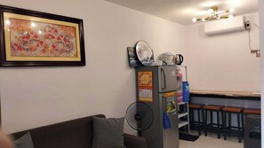 A living room with a couch, a refrigerator, a fan, a kitchen area with a counter and stools, and a framed picture on the wall.