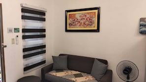 A room with a sofa, a framed painting, a window with blinds, a thermostat, and a wall-mounted fan.