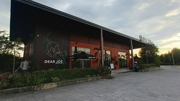 Een modern gebouw met een rode gevel, een groot bord waarop 'Dear Joe Coffee & Juice' staat en terrasstoelen met parasols.
