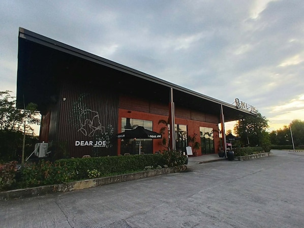 A modern building with a red facade, a large sign reading 'Dear Joe Coffee & Juice,' and outdoor seating with umbrellas.