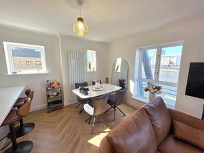 A modern living room with a dining area, large windows, a leather sofa, and a hanging light fixture. - Bright Modern 2BR Flat Sleeps 5 Royal Docks (Greater London)