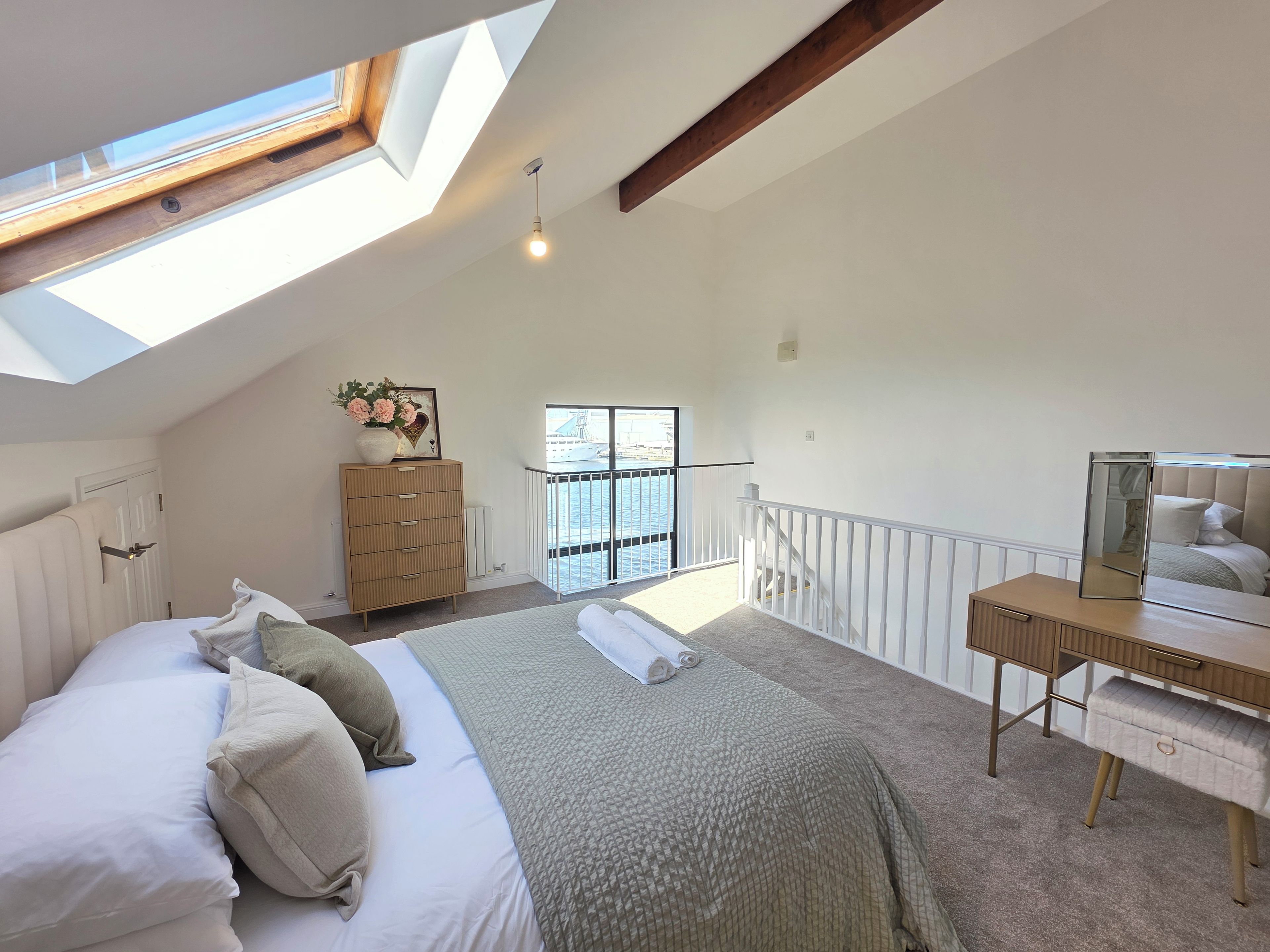 A bedroom with a skylight, a bed with pillows, a dresser, a desk with a mirror, and a view of the outside.