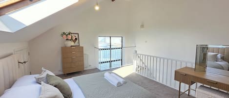 A bedroom with a skylight, a bed with pillows, a dresser, a desk with a mirror, and a view of the outside.