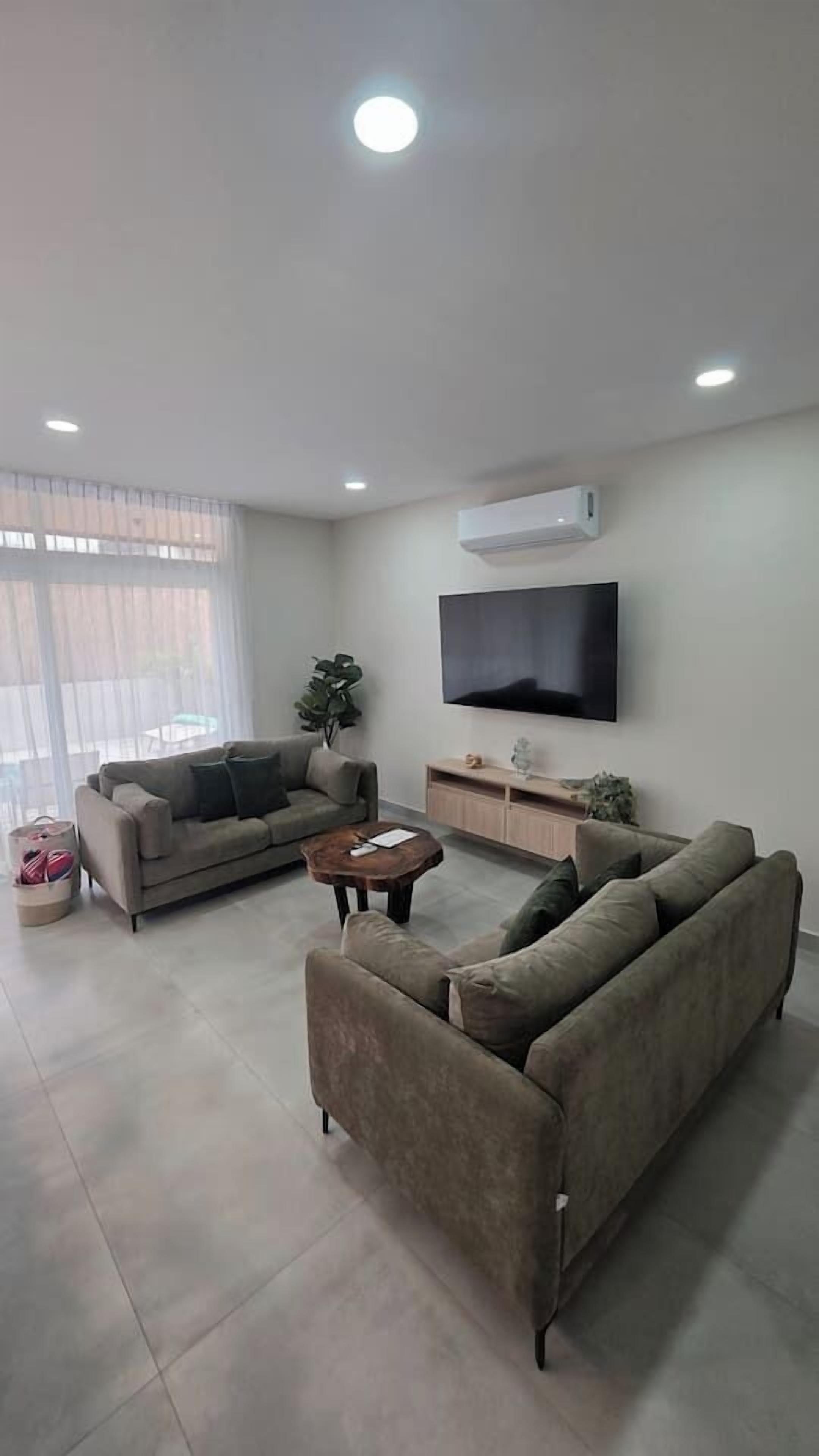 A modern living room with a flat-screen TV, two sofas, a coffee table, and a large window with sheer curtains.