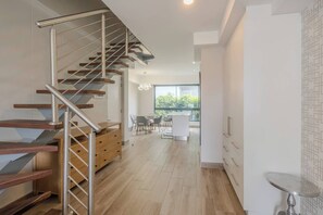 A modern interior with a wooden staircase, a dining area with chairs, and a view through a large window. - Luxury 2 BR Loft w Pool, Patio & Menlyn Maine (Pretoria)