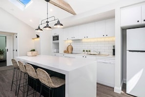 Cozinha moderna com armários brancos, uma ilha central com banquetas, iluminação pendente e forno embutido.