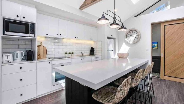 Una cocina moderna con armarios blancos, una isla central con taburetes y un microondas, además de un reloj en la pared.