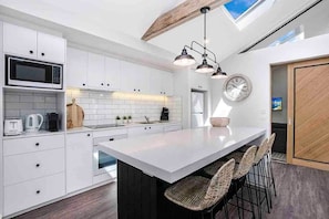 A modern kitchen with white cabinetry, a central island with bar stools, a microwave, and a clock on the wall. - Day Spa Apartment 1 & 2 in CBD (Port Fairy)