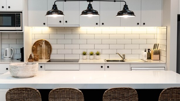 Une cuisine moderne avec des armoires blanches, un plan de travail blanc, un four à micro-ondes encastré, un évier et trois luminaires suspendus.