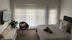 Una camera da letto con un letto, una sedia, un termosifone, uno specchio e tende leggere.
