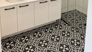 Un bagno con un mobile lavabo bianco, un lavandino rotondo, uno specchio, una finestra e una doccia dotata di un tubo nero, oltre a piastrelle decorative sul pavimento.