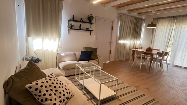 A living room with a sofa, a coffee table, a dining table with chairs, a wooden ceiling, and a door. - Large Guesthouse 80m in lovely Ramot (Ramot)