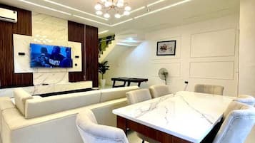 Un espace repas moderne avec une table en marbre, des chaises à revêtement blanc, un canapé et un téléviseur fixé au mur.