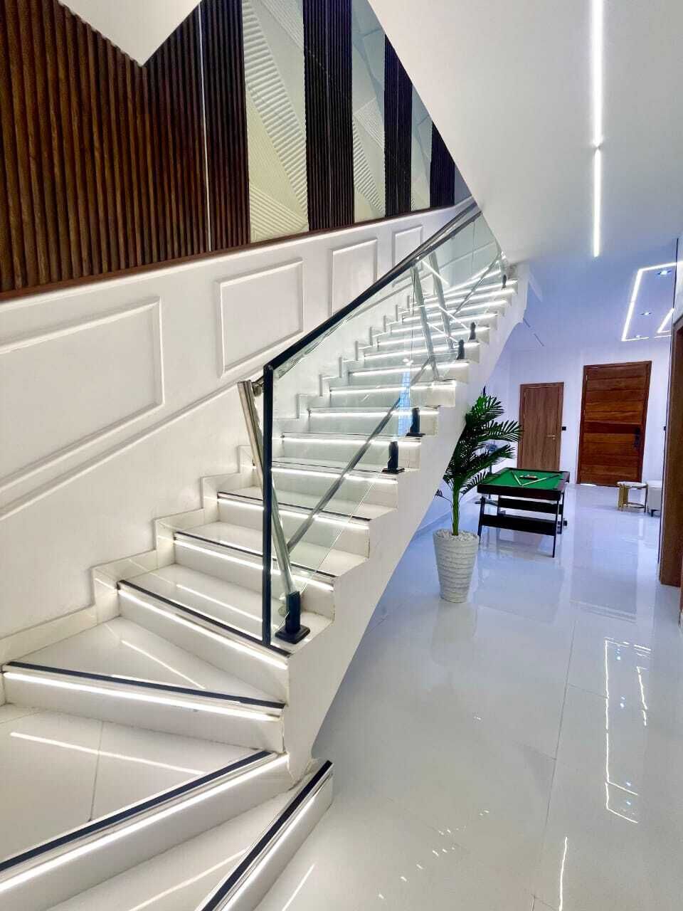Un escalier moderne avec des rampes en verre, une table de billard verte et une porte en bois, le tout dans un couloir lumineux et bien éclairé.